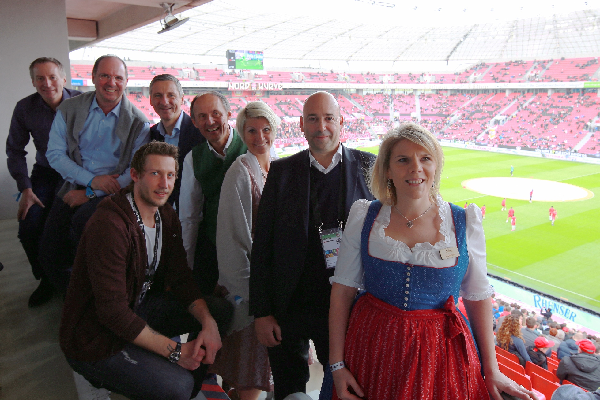 Eine Gruppe lächelnder Menschen in einer Stadionsuite, im Hintergrund ist ein Fußballfeld zu sehen.