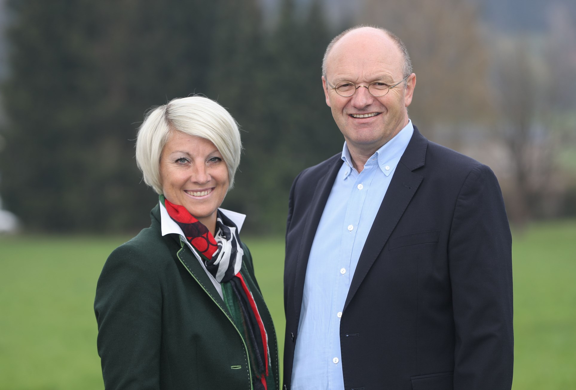Eine lächelnde Frau und ein lächelnder Mann stehen zusammen im Freien mit Grün und Bäumen im Hintergrund.