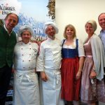 Sechs Erwachsene, darunter Köche und Frauen in österreichischer Tracht, posieren gemeinsam und lächeln vor einer Alpenkulisse.