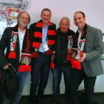 Vier lächelnde Männer mit Bayer-Leverkusen-Schals posieren gemeinsam in einer Halle vor Plakaten an der Wand.