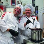 Zwei lächelnde Köche in weißen Uniformen und Mützen bereiten an einem Tresen mit einem großen Topf Schokoladenleckereien zu.