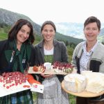 Drei Frauen im Freien in den Bergen halten Käseteller und Auszeichnungen und lächeln in die Kamera.