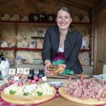 Lächelnde Frau an einem Marktstand, die Lebensmittel anbietet, mit Käse, Fleisch und Gläsern vor sich.