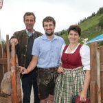 Drei Erwachsene in traditioneller österreichischer Kleidung stehen im Freien an einem Holzzaun, hinter ihnen liegen grüne Hügel.