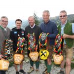 Fünf Männer im Freien, lächelnd und mit geschmückten Kuhglocken und Helmen in der Hand, Berge im Hintergrund.