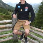 Ein Mann in Shorts und Jacke sitzt auf einem Holzzaun in einer bergigen, grasbewachsenen Landschaft.