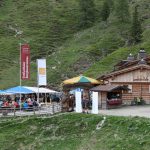 Menschen, die unter freiem Himmel in einem Berghüttencafé mit Sonnenschirmen speisen, umgeben von grasbewachsenen Hügeln und Bäumen.