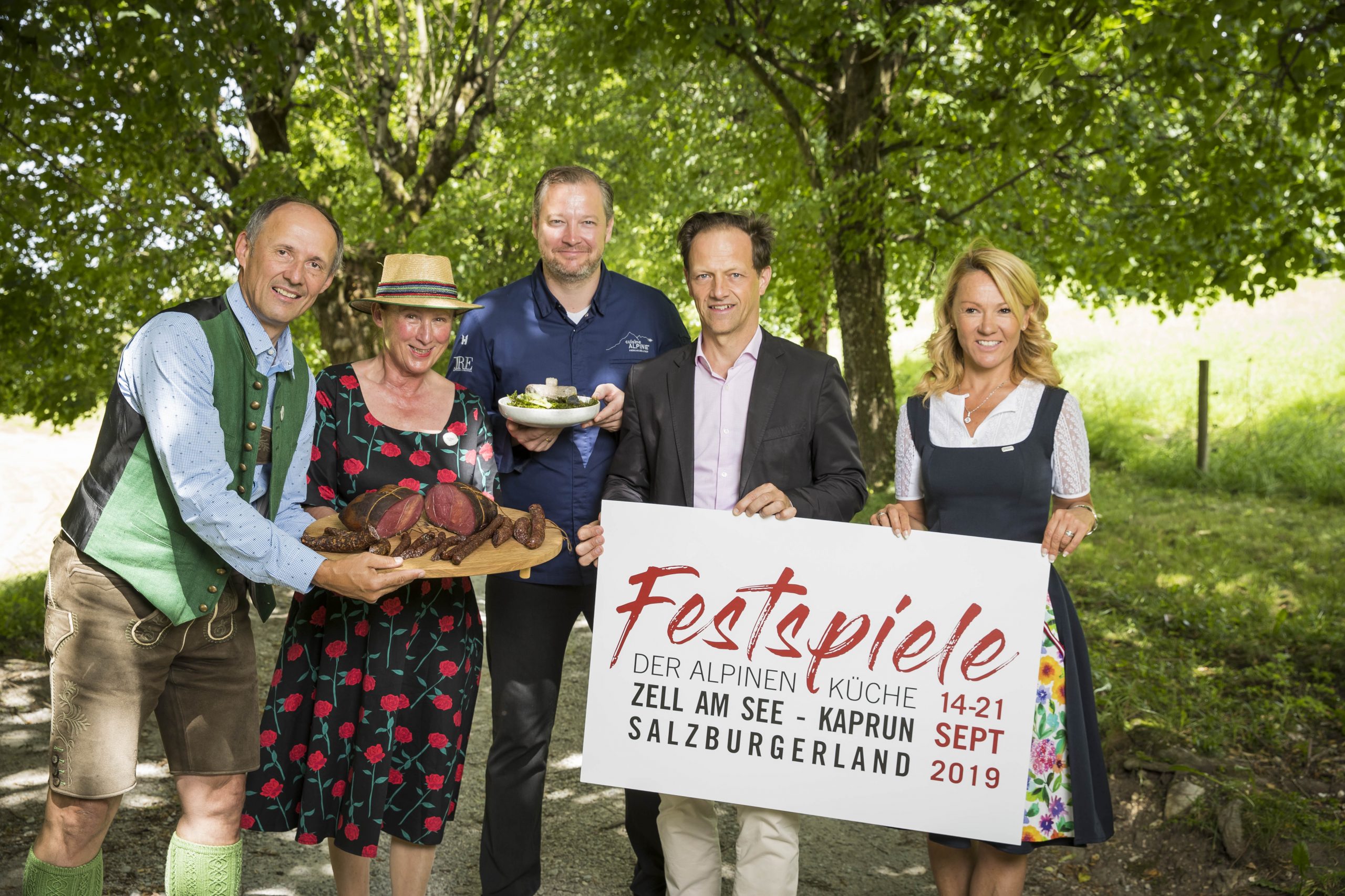 Fünf Personen in österreichischer Tracht halten an einem sonnigen Tag im Freien unter Bäumen Essen und ein Festschild in der Hand.