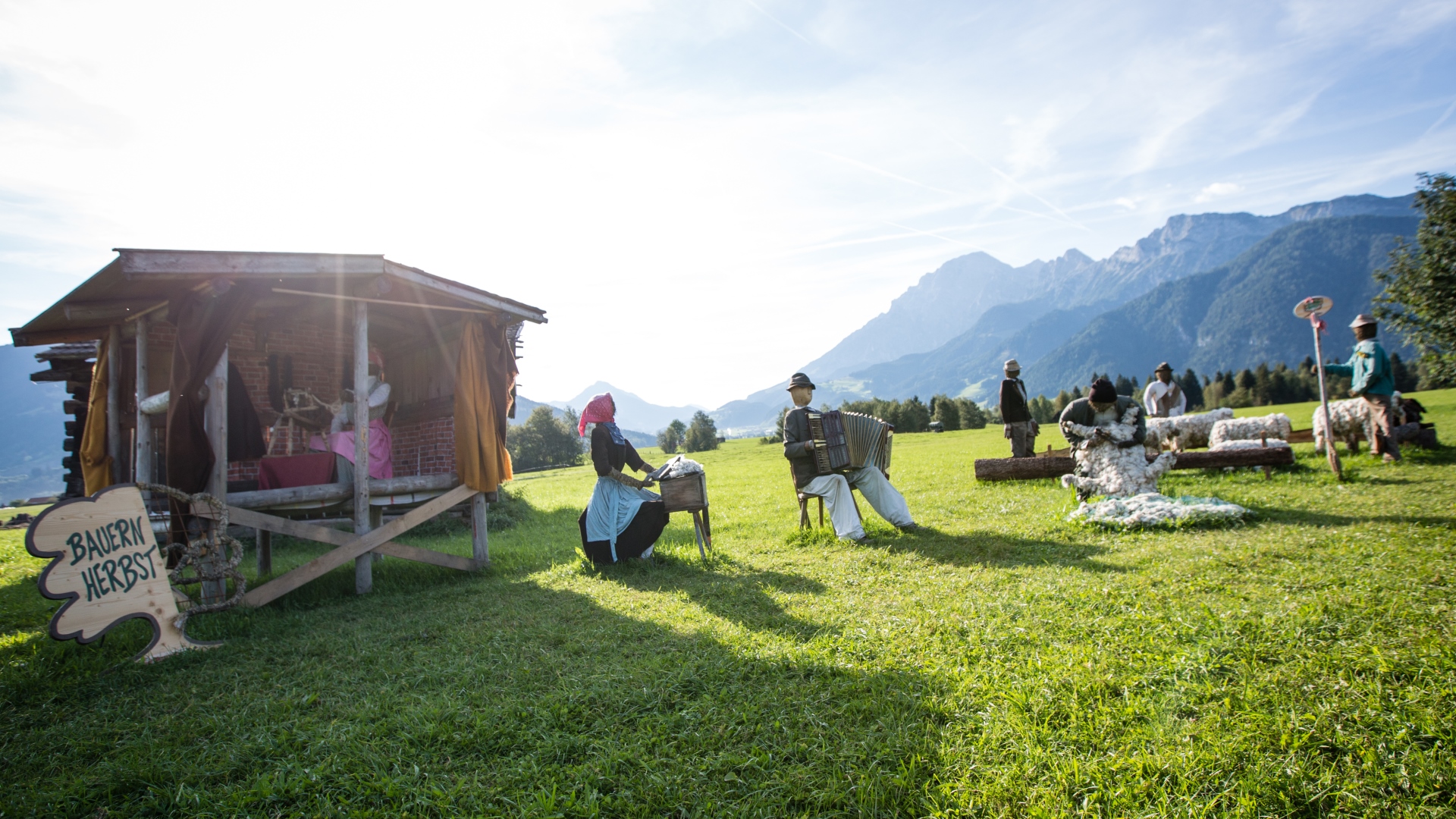 Menschen in traditioneller Kleidung sitzen neben einer Holzhütte auf einer sonnigen Bergwiese mit einem Bauernherbst-Schild.