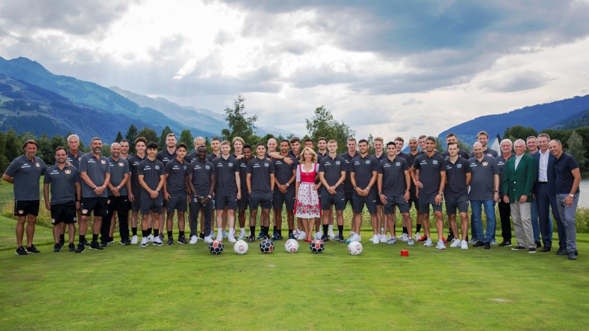 Eine große Fußballmannschaft posiert im Freien auf einer Wiese mit Bergen im Hintergrund, in der Mitte steht eine Frau in einem Kleid.