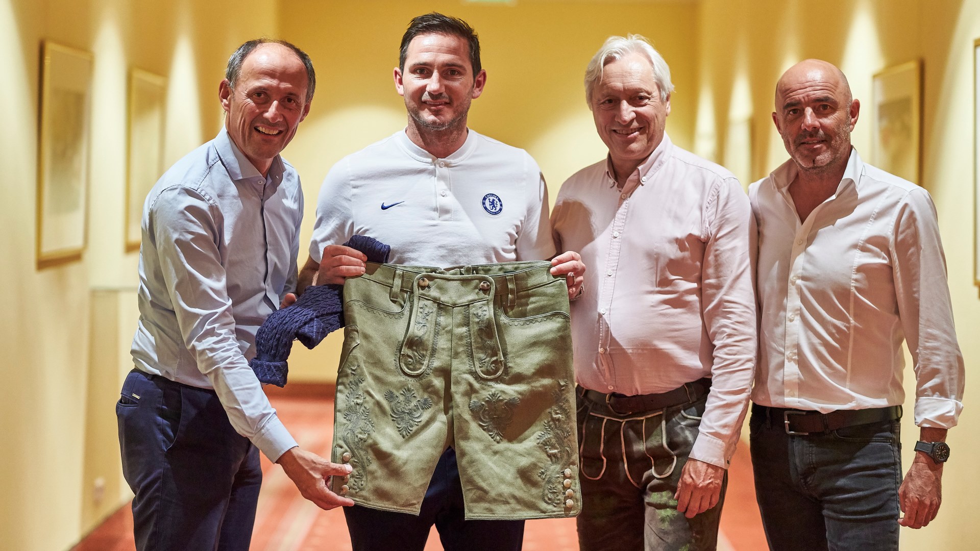 Vier Männer stehen in einem Hausflur, einer von ihnen trägt eine traditionelle grüne österreichische Lederhose und lächelt in die Kamera.