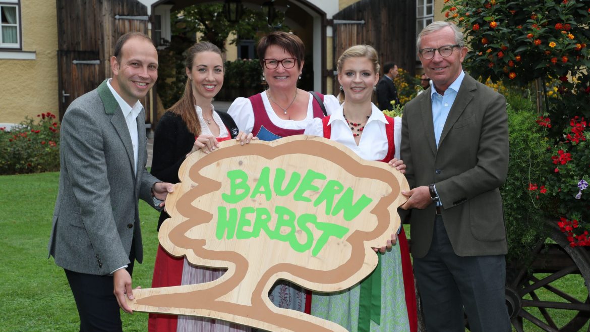 Fünf Personen in traditioneller Kleidung halten ein hölzernes Bauernherbst-Schild und lächeln im Freien neben einem Gebäude und Blumen.