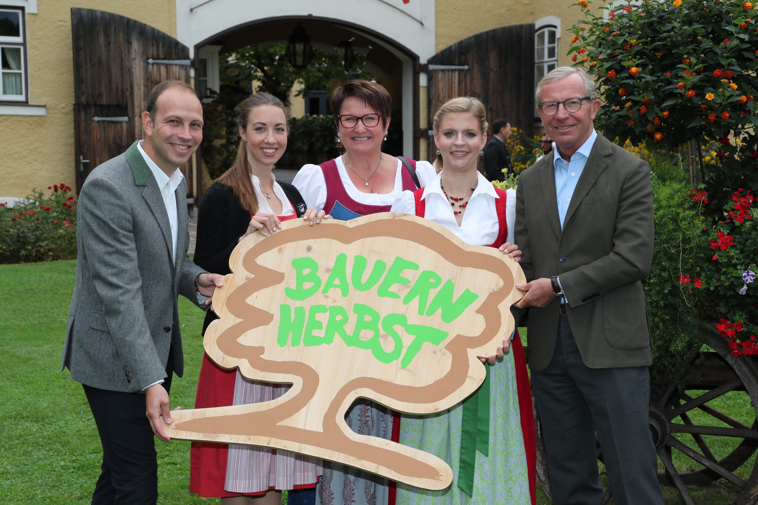 Fünf Personen in traditioneller Kleidung halten ein hölzernes Bauernherbst-Schild und lächeln im Freien neben einem Gebäude und Blumen.