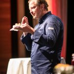 Ein Koch in einer blauen Jacke spricht leidenschaftlich auf der Bühne, in der Hand einen kleinen Gegenstand und eine Serviette.