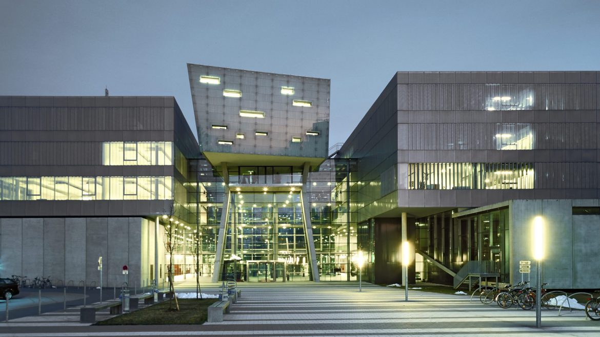 Modernes Gebäude aus Glas und Beton mit geometrischer Architektur, das in der Abenddämmerung beleuchtet wird, vor dem Fahrräder geparkt sind.