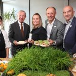 Fünf Erwachsene posieren in einem Haus mit Lebensmitteln und Grünzeug auf einem Tisch; eine Frau trägt eine Kochuniform und eine Mütze.