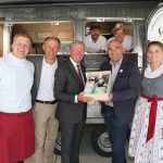 Sechs Personen lächeln vor einem SalzburgLand-Imbisswagen, eine hält einen Geschenkkorb mit regionalen Produkten.