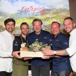 Fünf Köche lächeln und halten eine Holzkiste mit Kräutern bei einer kulinarischen Veranstaltung vor einer malerischen Kulisse.