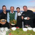 Vier lächelnde Männer stehen bei Sonnenuntergang im Freien hinter einem Tisch mit Speisen und halten Schüsseln mit Zutaten.