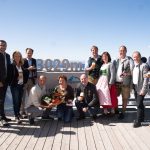 Eine Gruppe von Menschen posiert mit Getränken und einem Obstkorb auf einer Bergterrasse in 3029 Metern Höhe unter einem klaren Himmel.