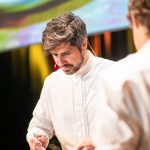 Ein Koch im weißen Kittel konzentriert sich bei einer hell erleuchteten Veranstaltung auf das Ausgießen von Soße aus einer Pfanne.