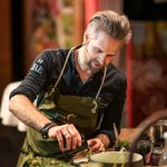 Ein Mann mit Schürze und Headset bereitet an einer Küchentheke mit frischen Kräutern und Zutaten Speisen zu.