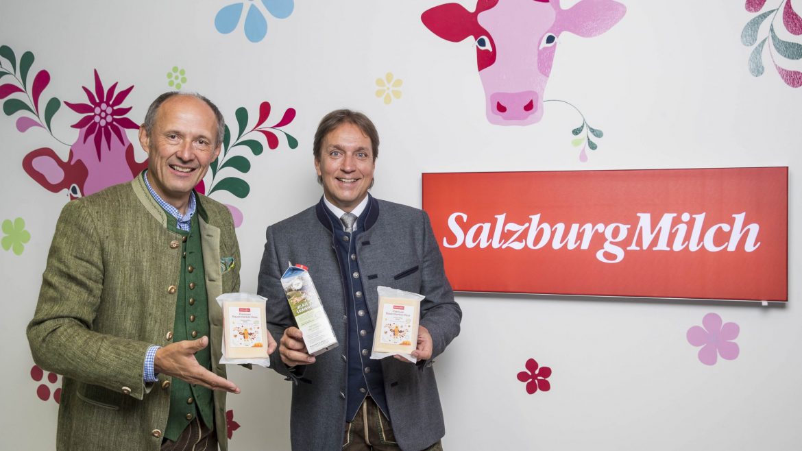 Zwei Männer in traditioneller österreichischer Kleidung halten SalzburgMilch-Milchprodukte in der Hand und stehen neben einem roten SalzburgMilch-Schild.