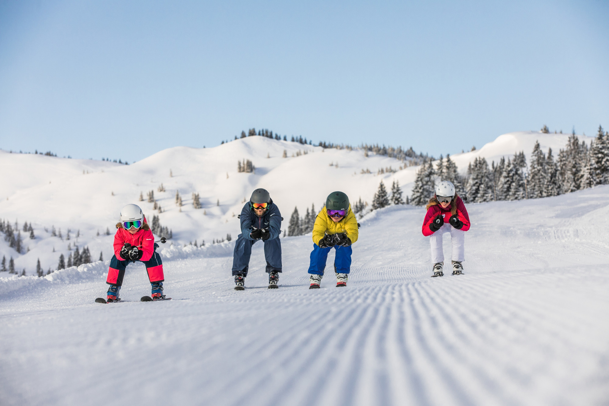 Vier Kinder in bunten Skiklamotten fahren auf einem sonnigen, verschneiten Berg mit Bäumen im Hintergrund bergab.