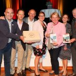 Sieben lächelnde Menschen auf der Bühne, die Broschüren und eine Holzkiste in der Hand halten, bei einer Indoor-Veranstaltung mit warmer Beleuchtung.