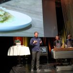 Ein Koch präsentiert sich auf der Bühne mit drei Assistenten, hinter denen ein großes Lebensmittelbild angezeigt wird.