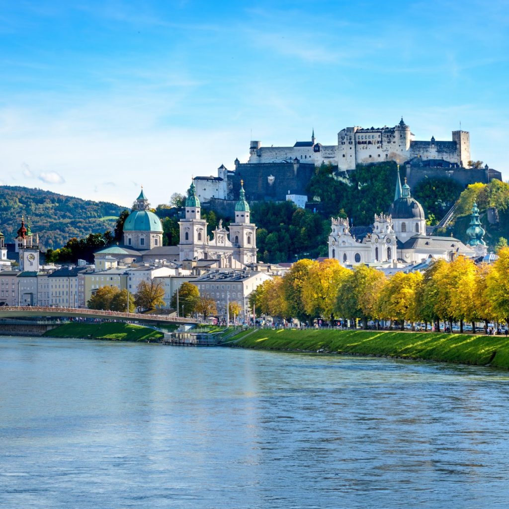 Ein Fluss fließt durch Salzburg, Österreich, mit historischen Gebäuden und einer Festung auf einem Hügel unter einem blauen Himmel.