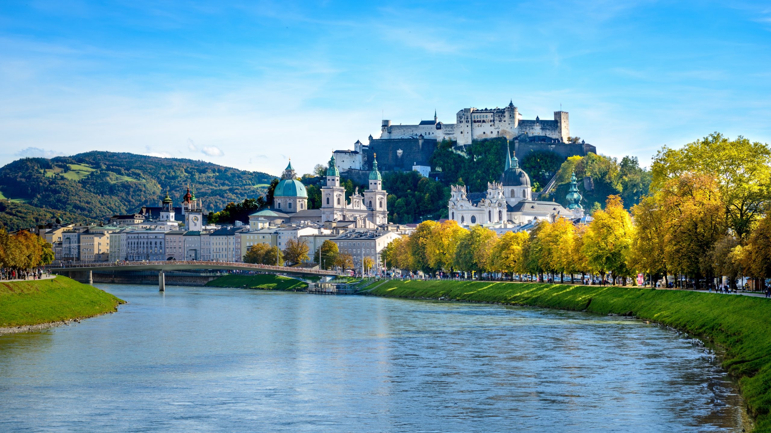 Ein Blick auf das Flussufer von Salzburg, Österreich, mit der Festung Hohensalzburg auf einem Hügel und Herbstbäumen.