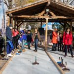 Menschen spielen einen Wintersport auf einem Platz im Freien, umgeben von Zuschauern unter einem Holzdach.