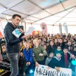 Ein Mann spricht zu einer Menge aufgeregter Kinder in einem großen Veranstaltungszelt mit bunten Bannern.