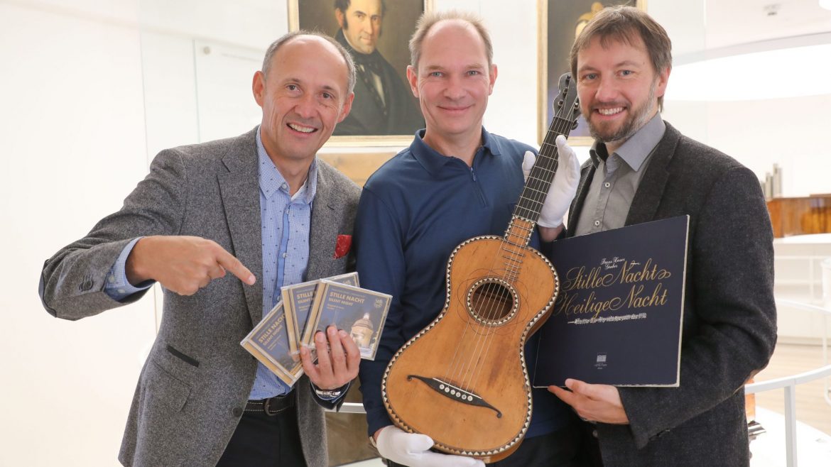 Drei lächelnde Männer posieren mit einer Gitarre, CDs und einer Schallplatte vor gerahmten Porträts.