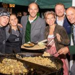 Sechs lächelnde Menschen posieren bei einer Veranstaltung im Freien neben einer großen Pfanne mit Speisen, einige halten Teller und Servierutensilien.