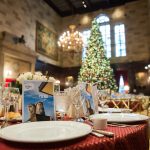 Eleganter Esstisch für eine Mahlzeit in einer großen Halle mit einem geschmückten Weihnachtsbaum im Hintergrund.