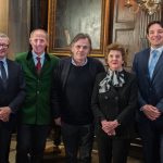 Fünf formell gekleidete Personen stehen in einem Innenraum zusammen und lächeln für ein Gruppenfoto vor einer holzgetäfelten Wand.