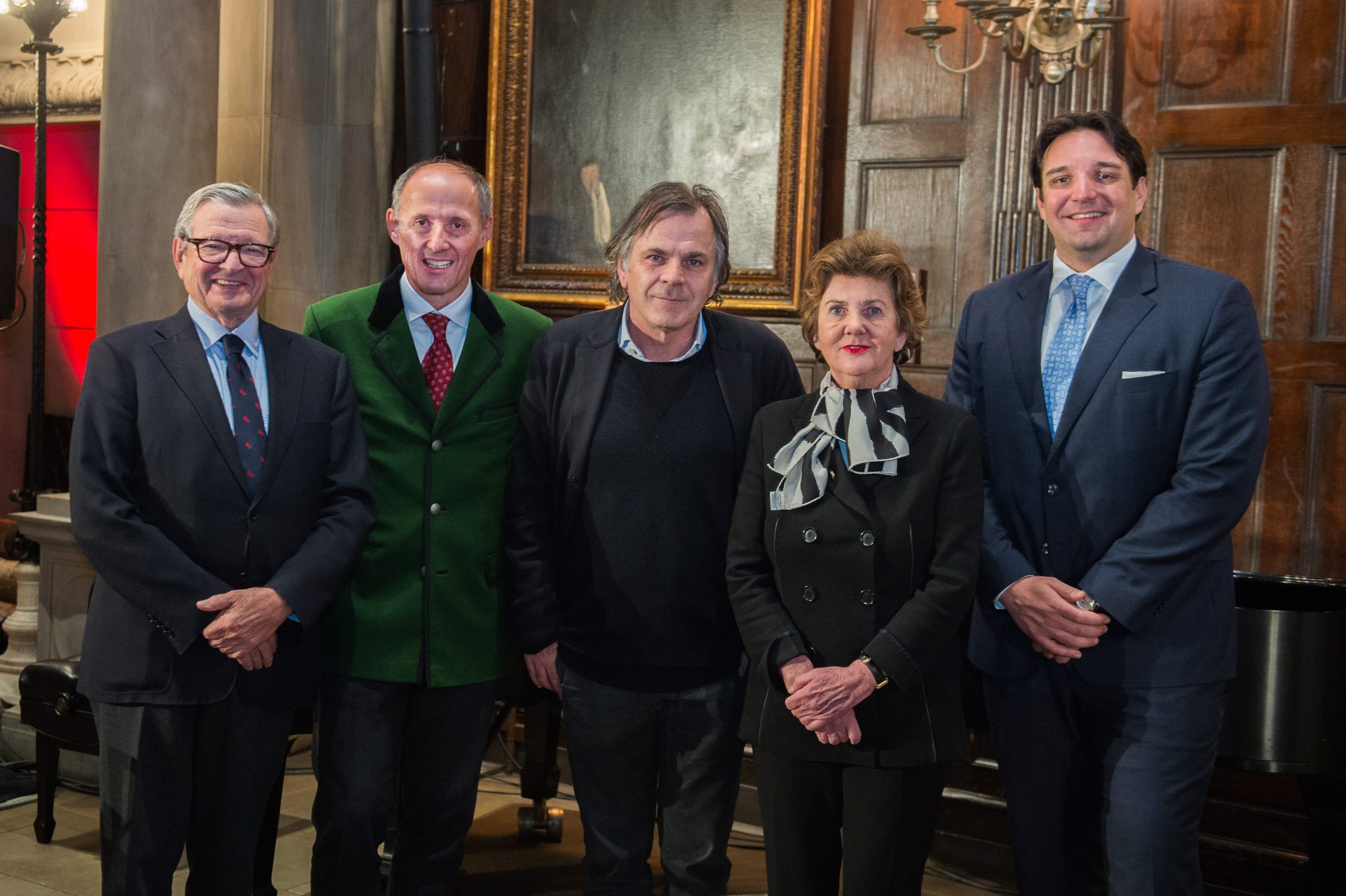 Fünf formell gekleidete Personen stehen in einem Innenraum zusammen und lächeln für ein Gruppenfoto vor einer holzgetäfelten Wand.