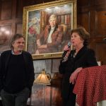 Eine Frau spricht in ein Mikrofon neben einem Mann, im Hintergrund ist ein Porträtgemälde zu sehen.