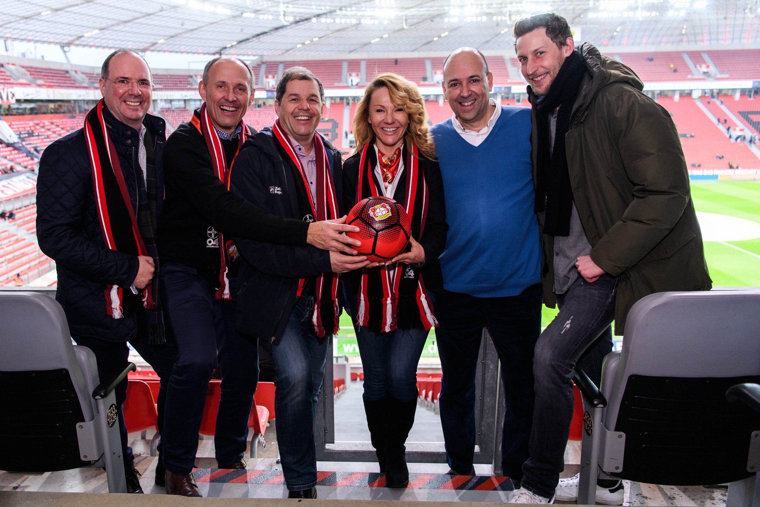 Sechs lächelnde Menschen mit Schals posieren zusammen und halten einen Fußball in einem Stadion mit Blick auf das Spielfeld.