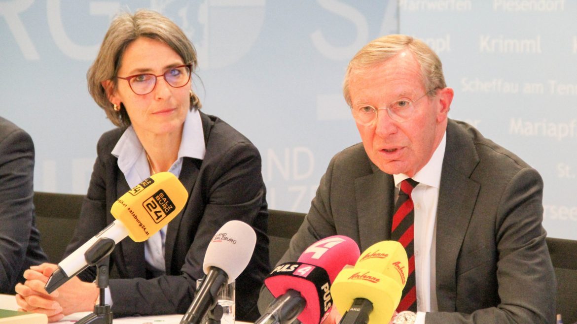 Eine Frau und ein Mann in Geschäftskleidung sprechen auf einer Pressekonferenz mit mehreren Mikrofonen vor ihnen.
