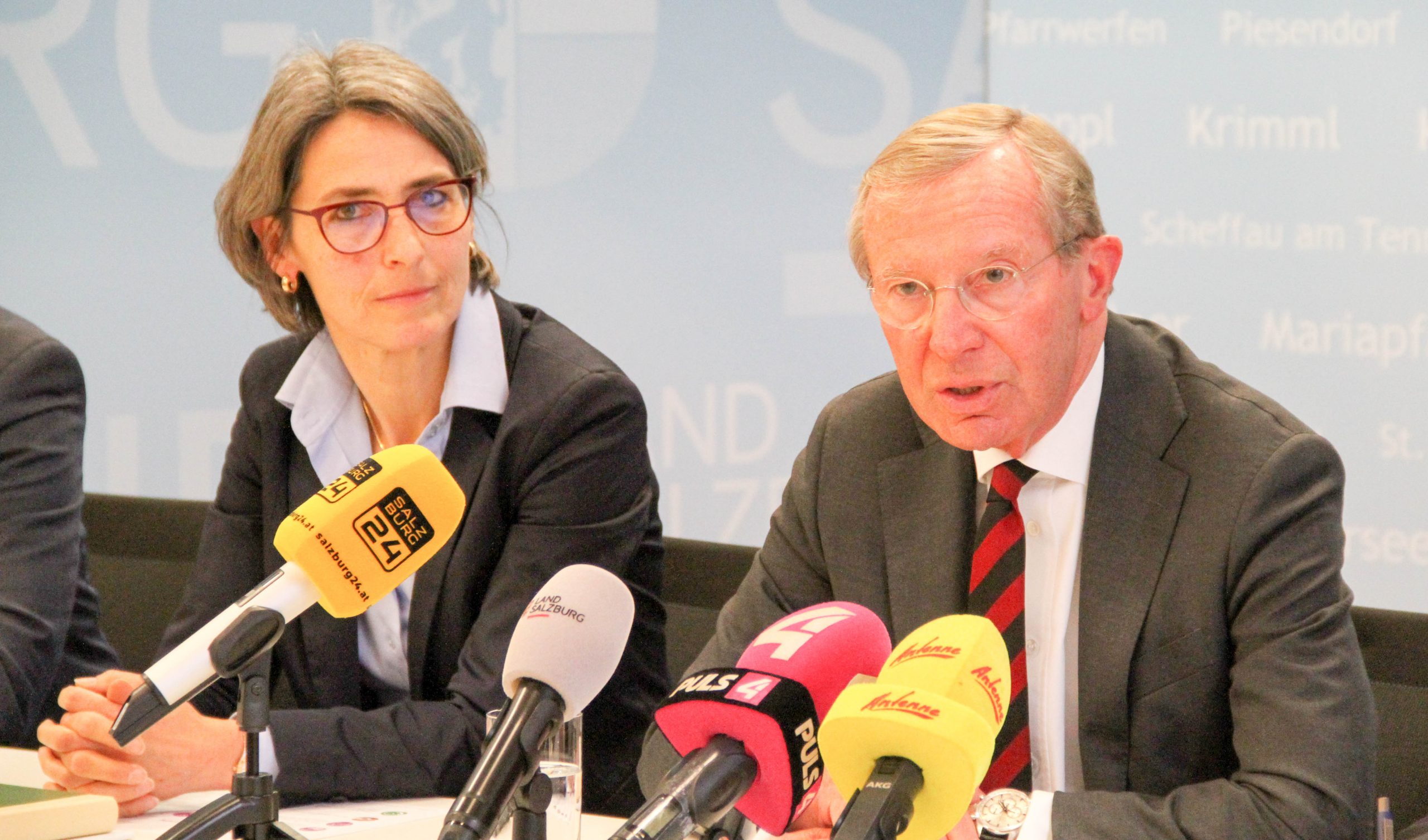 Eine Frau und ein Mann in Geschäftskleidung sprechen auf einer Pressekonferenz mit mehreren Mikrofonen vor ihnen.