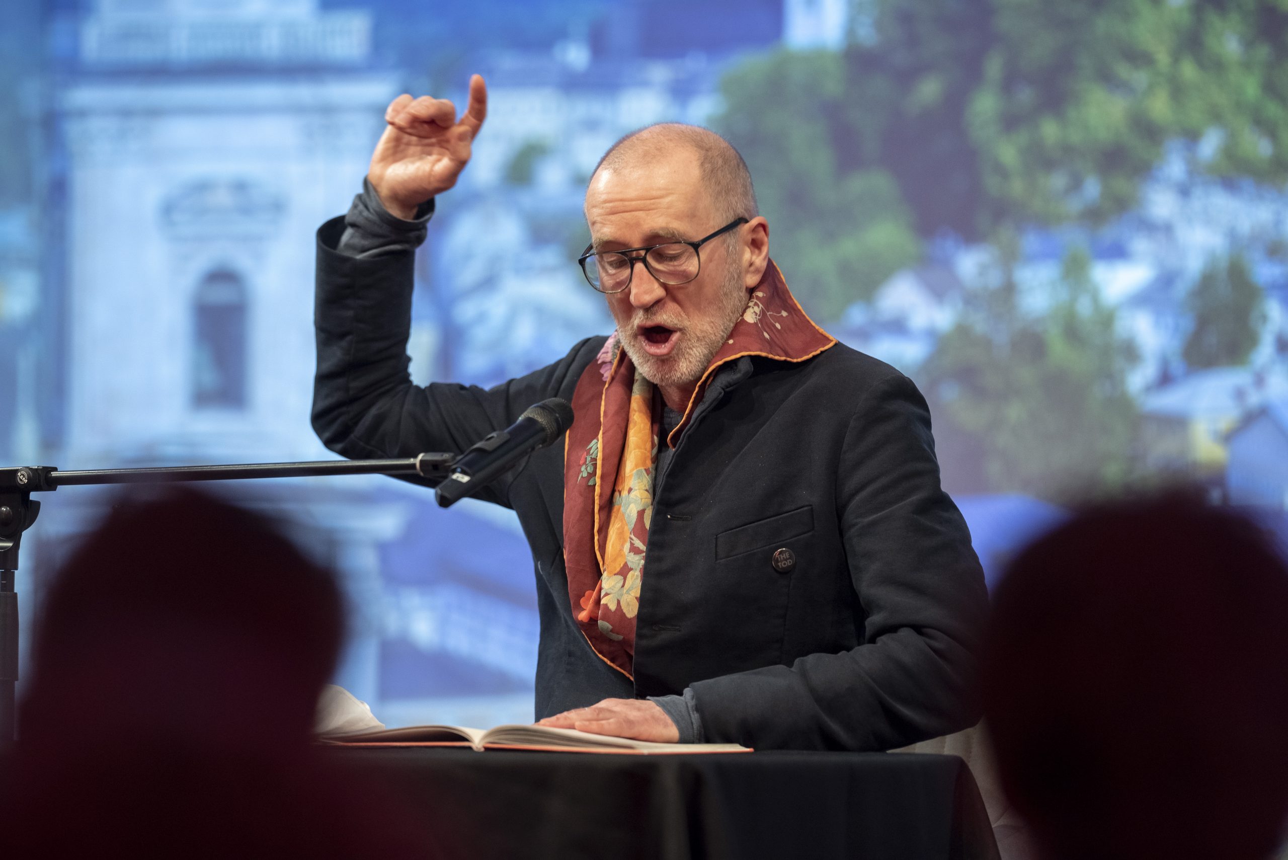 Ein Mann mit Brille liest leidenschaftlich in ein Mikrofon und hebt eine Hand zur Betonung.