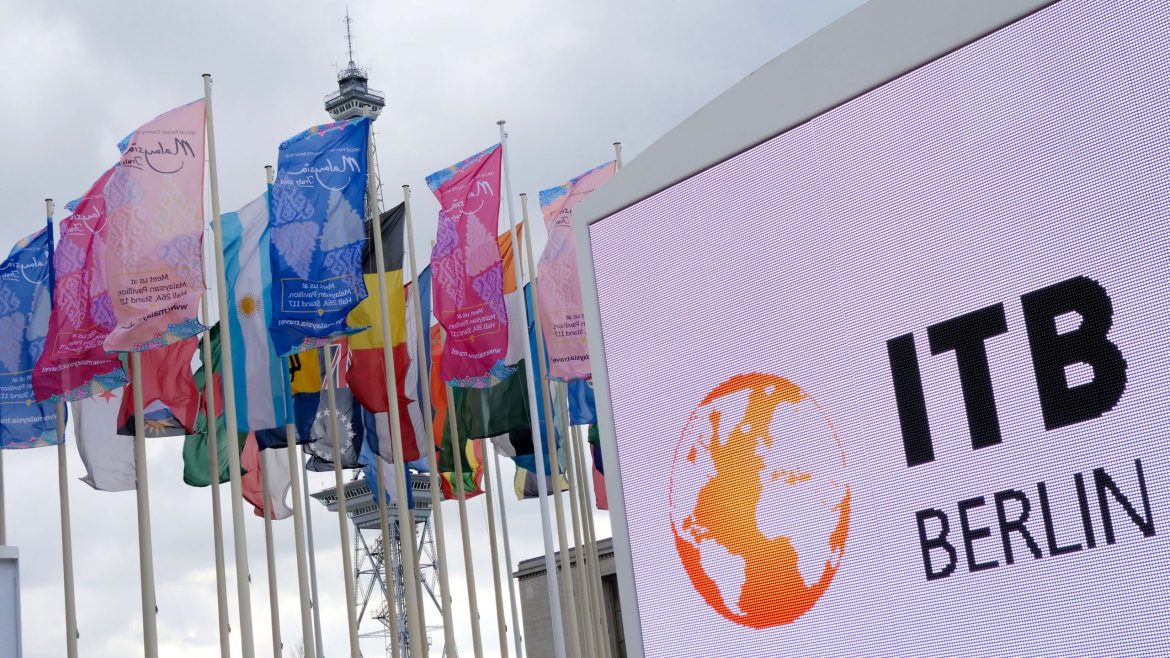 Bunte internationale Flaggen wehen neben einem großen ITB Berlin-Schild und einem Kommunikationsturm unter einem bewölkten Himmel.