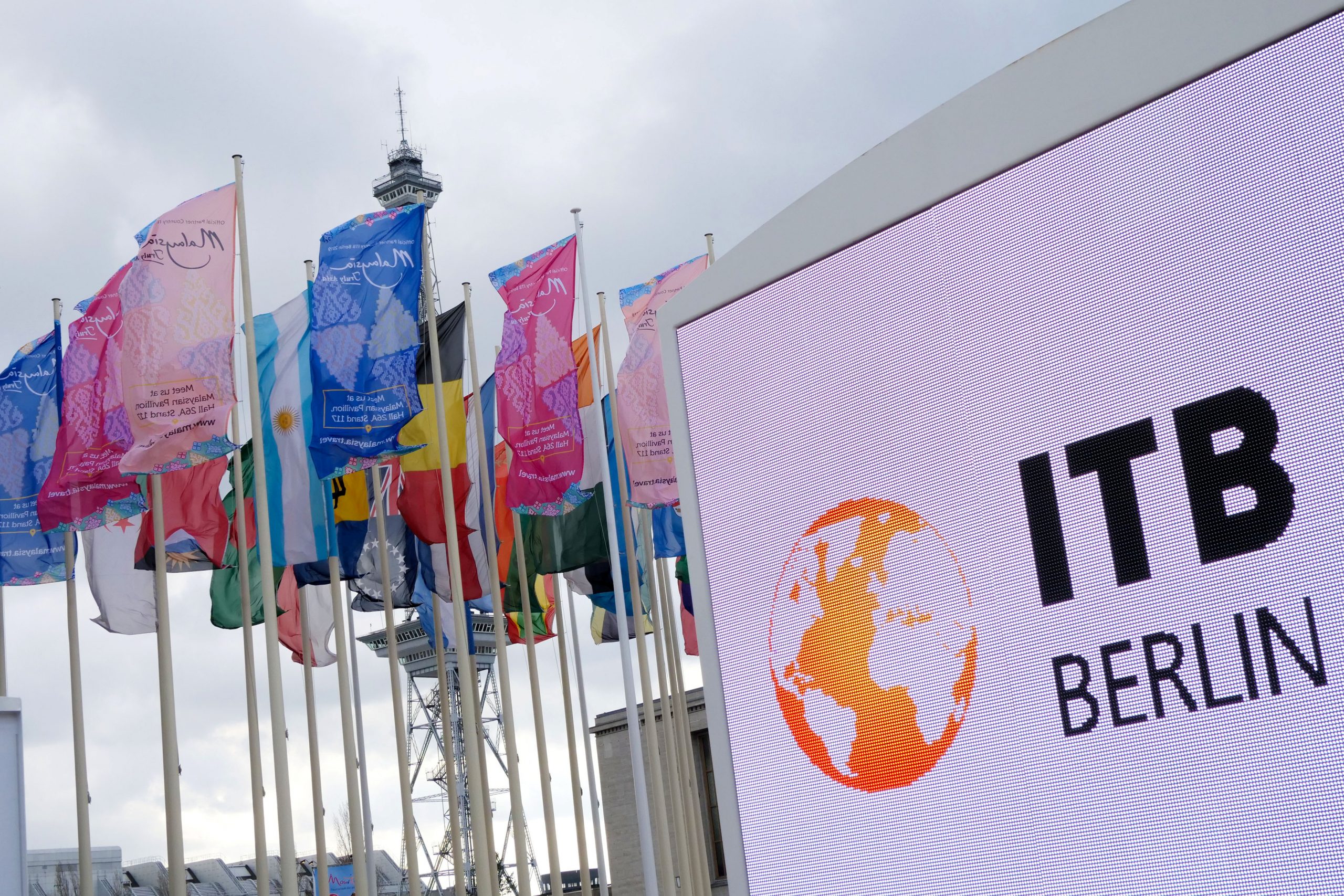 Bunte internationale Flaggen wehen neben einem großen ITB Berlin-Schild und einem Kommunikationsturm unter einem bewölkten Himmel.