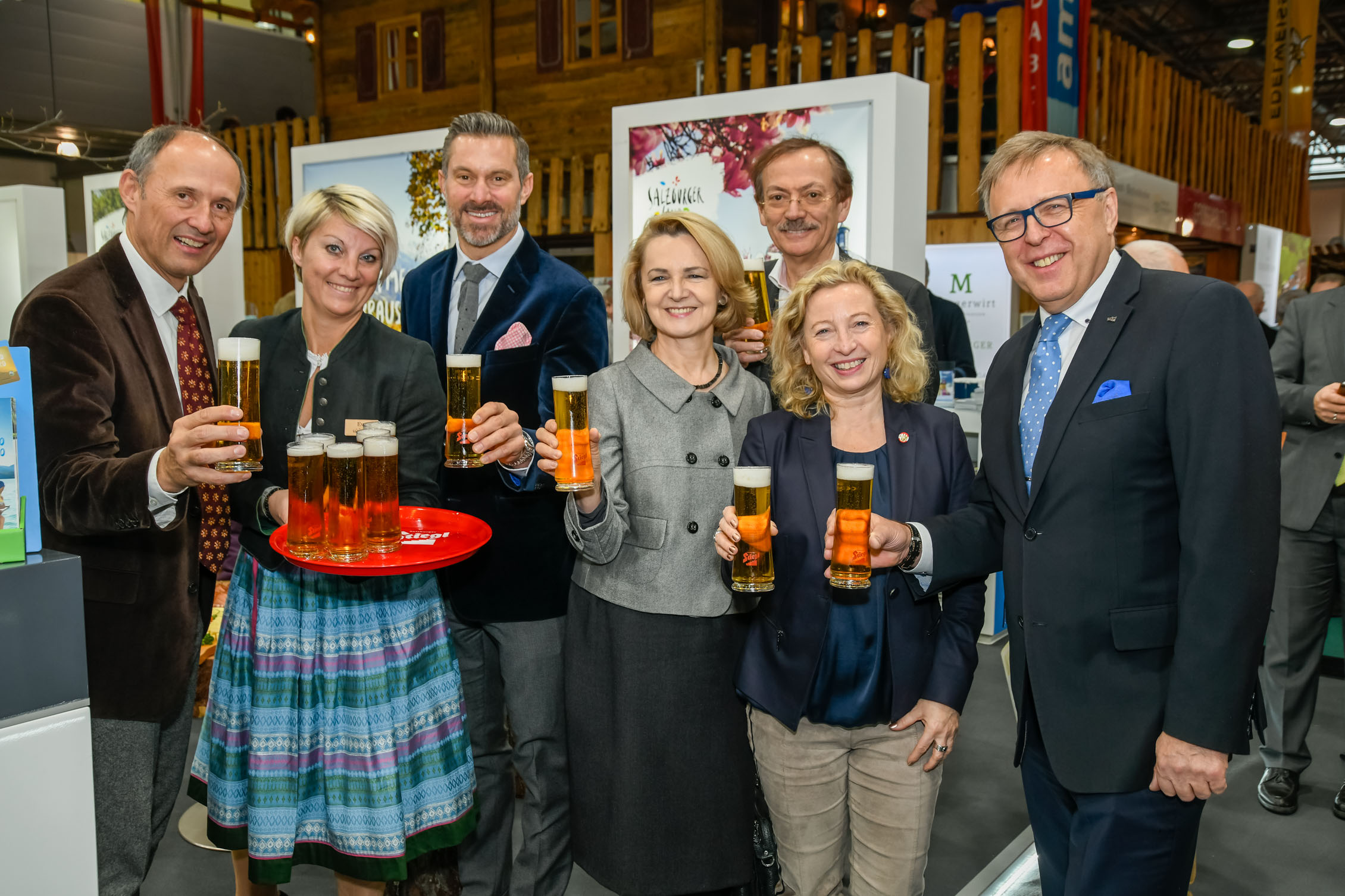 Sieben Personen in Geschäftskleidung lächeln und halten Biergläser bei einer Indoor-Veranstaltung.