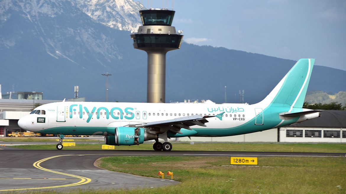 Ein Flynas-Flugzeug rollt auf einer Landebahn mit einem Kontrollturm und Bergen im Hintergrund.