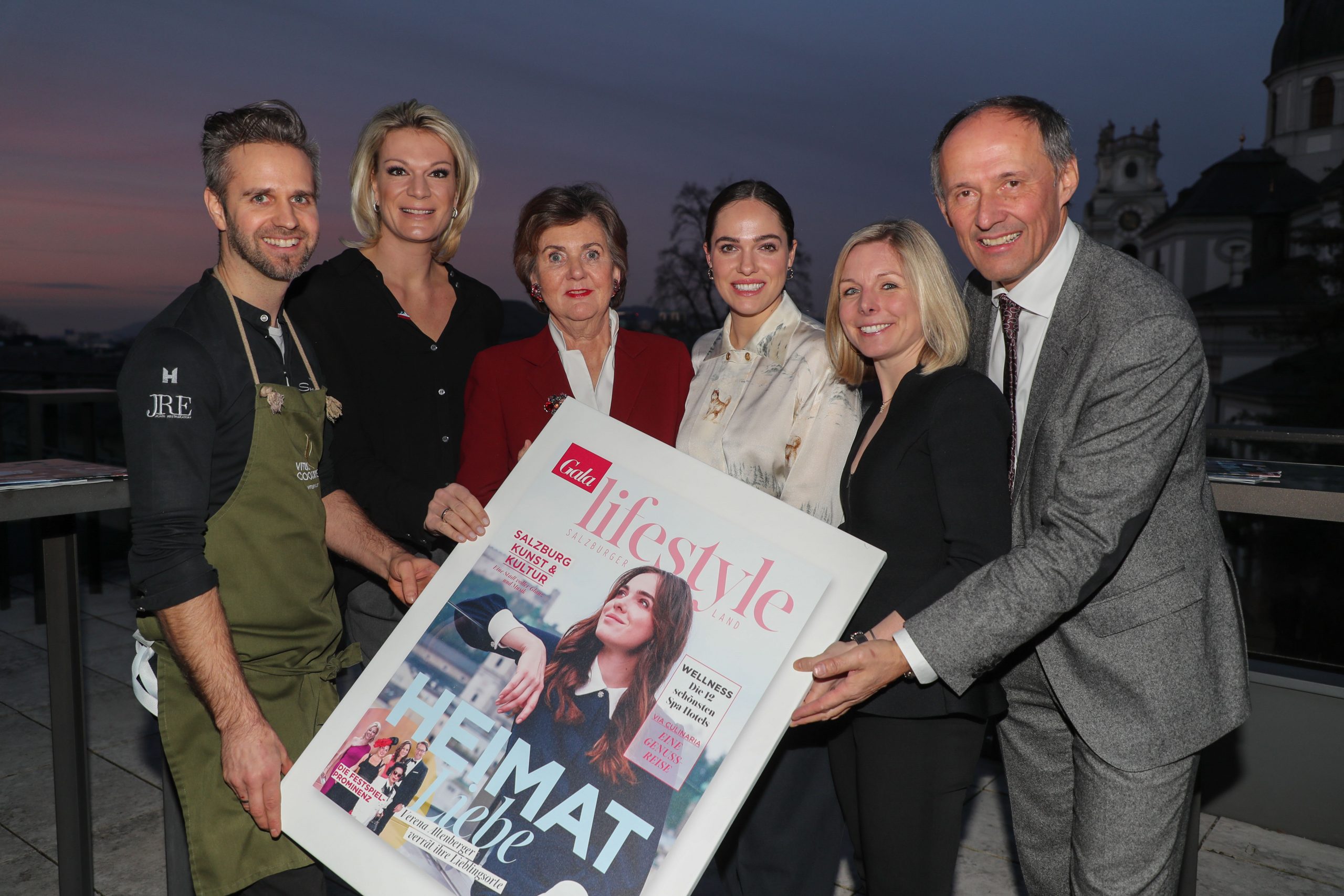 Sechs Personen posieren bei Sonnenuntergang im Freien und halten ein großes Lifestyle-Magazin, auf dem eine Frau abgebildet ist.