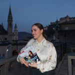 Eine Frau lächelt in der Abenddämmerung auf einem Balkon, in der Hand eine Zeitschrift, mit einer Stadtlandschaft und Kirchtürmen im Hintergrund.